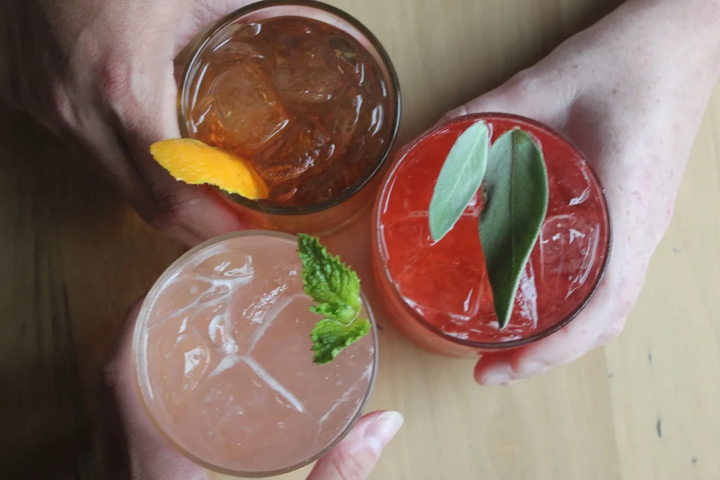 Overhead: hands holding three colorful cocktails with fresh garnishes.