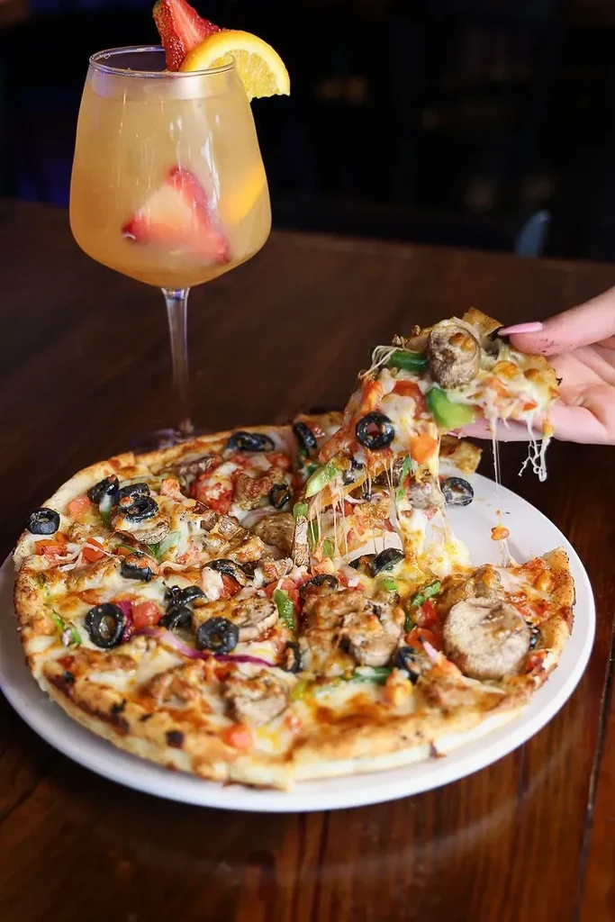 Hand lifts a pizza slice with cheese pull, varied toppings, and a cocktail.