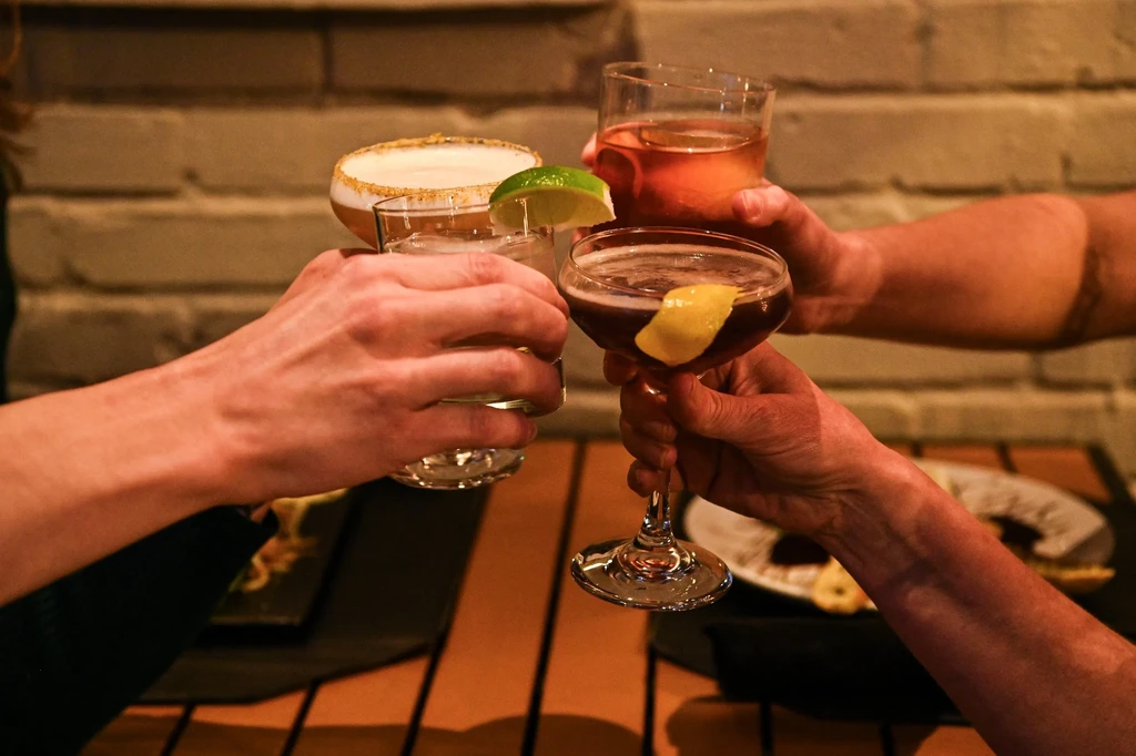 Four hands toast with diverse, colorful cocktails in a bar setting.