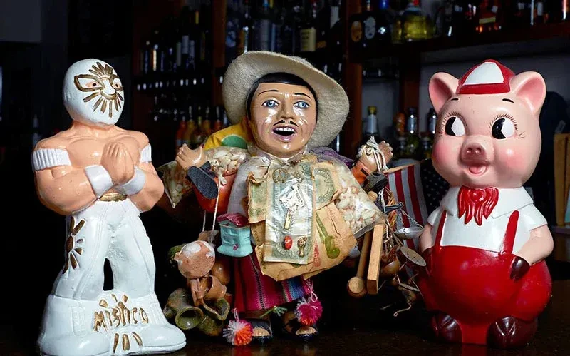 Three figurines: a luchador, traditional man, and pig, on a bar.
