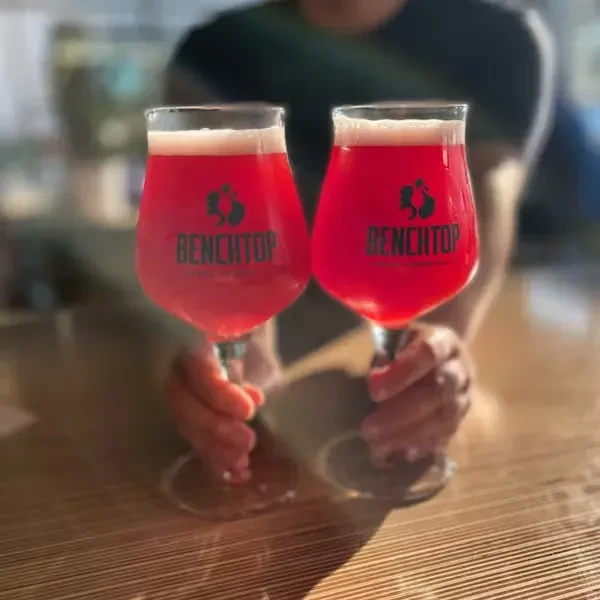 Two hands hold stemmed glasses of frothy pink Benchtop drinks.