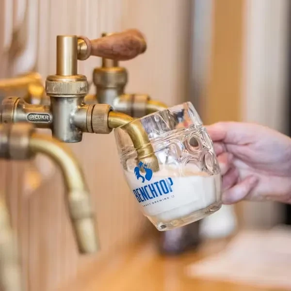 Hand pours frothy drink from brass tap into Benchtop Craft Brewing mug.