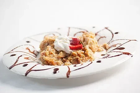 Warm fruit crisp, whipped cream, strawberry, chocolate drizzle on white plate.