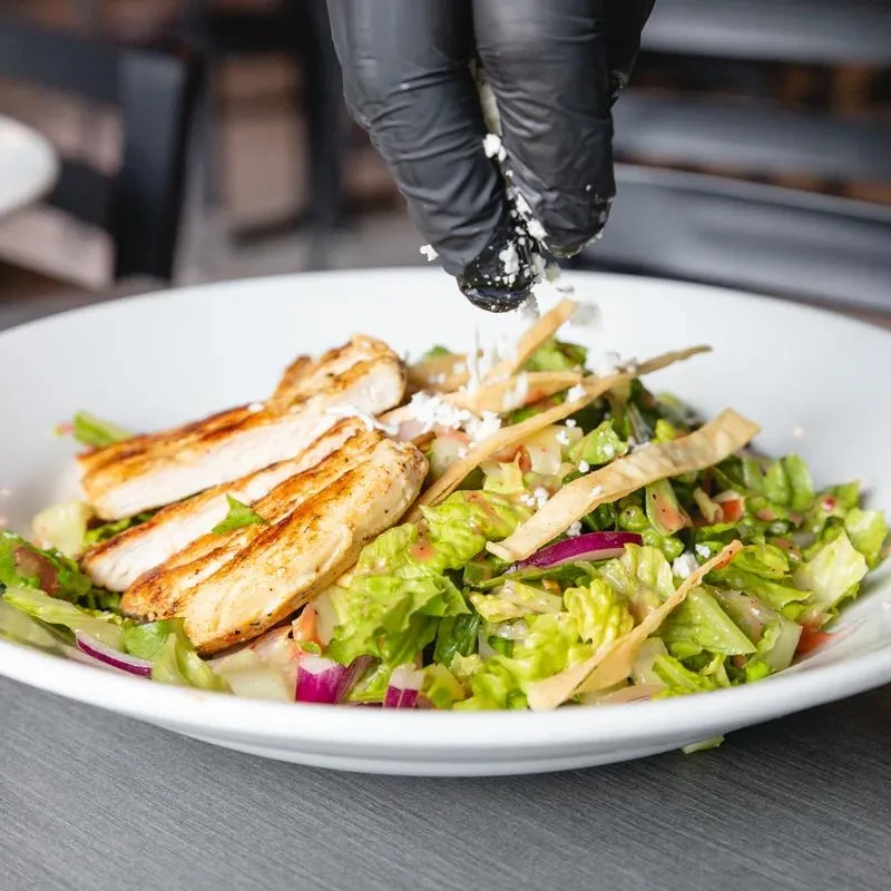 A gloved hand sprinkles cheese on a fresh salad with grilled chicken.