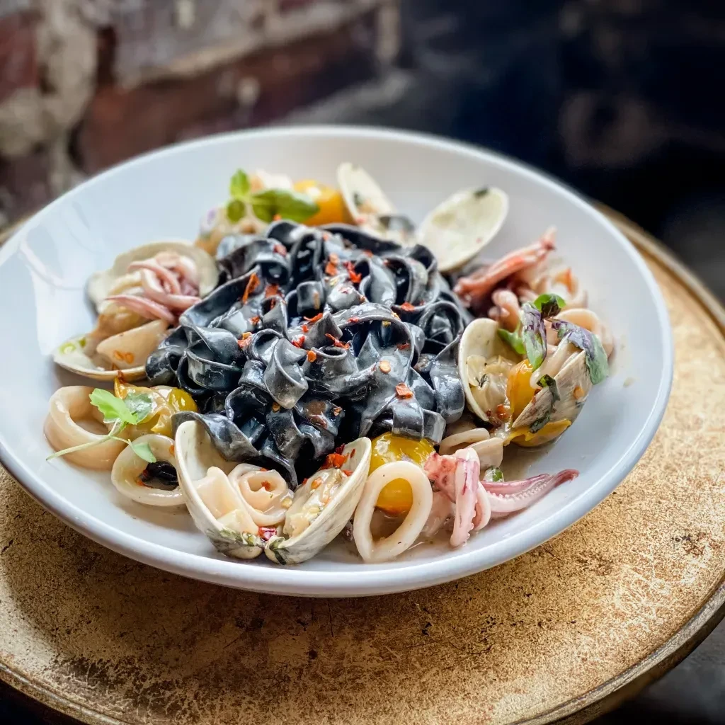 Close-up top-down: squid ink pasta with clams, squid, tomatoes, herbs.