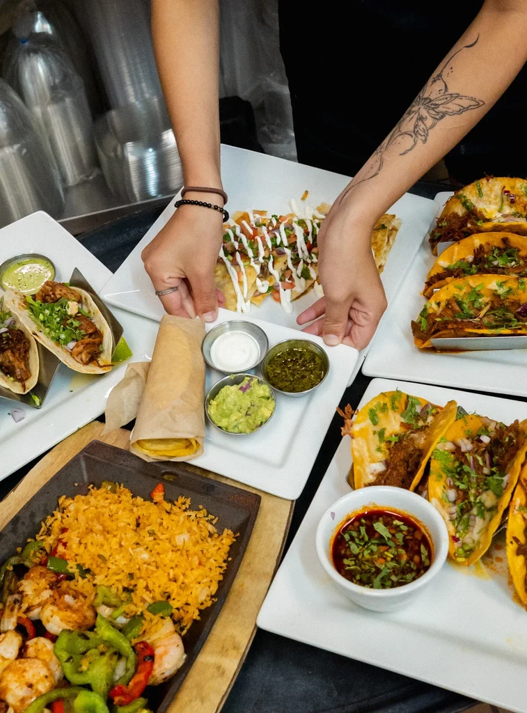 Hands arranging plates of Mexican tacos, nachos, and shrimp.