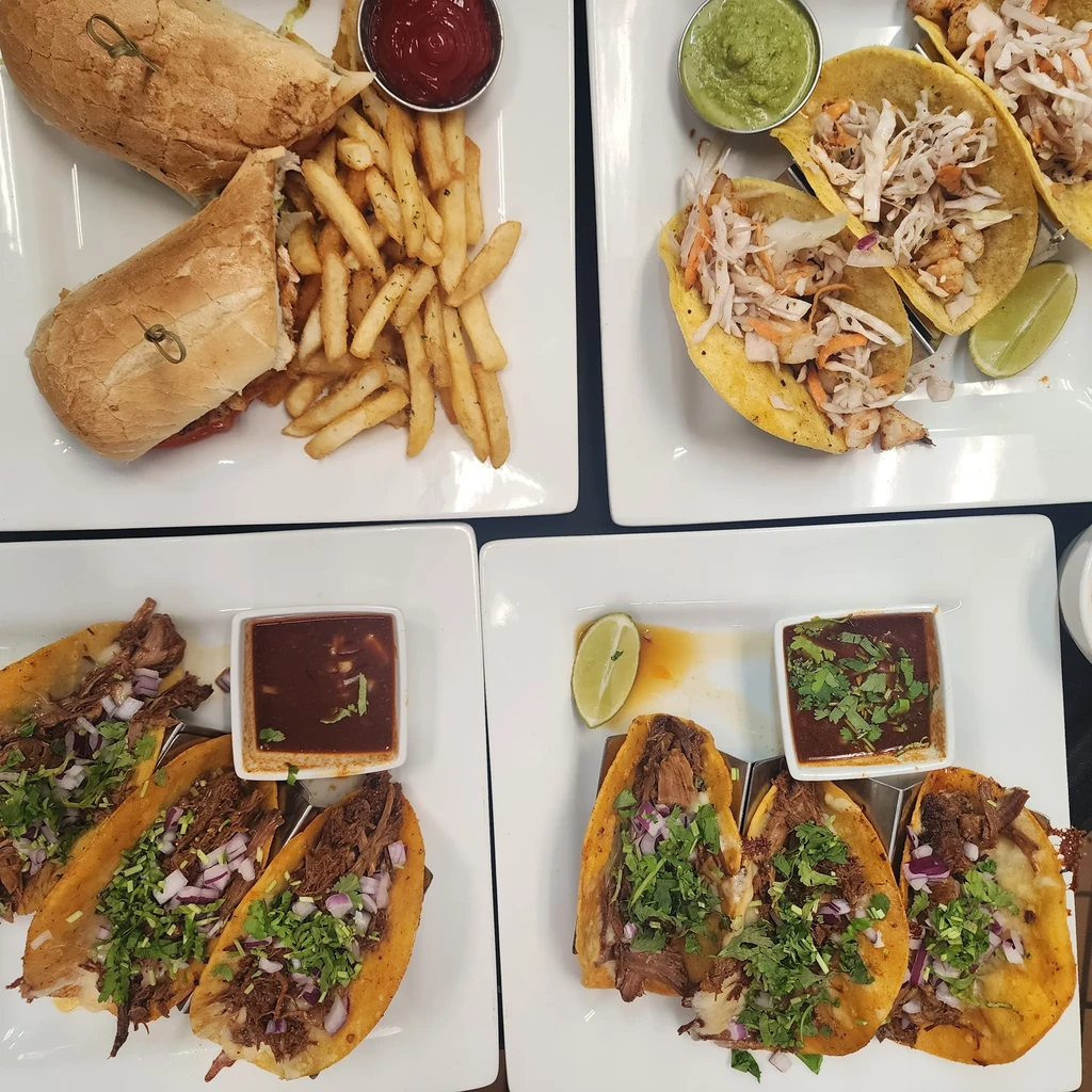 Overhead view of four distinct platters: sandwich, shrimp, and birria tacos.
