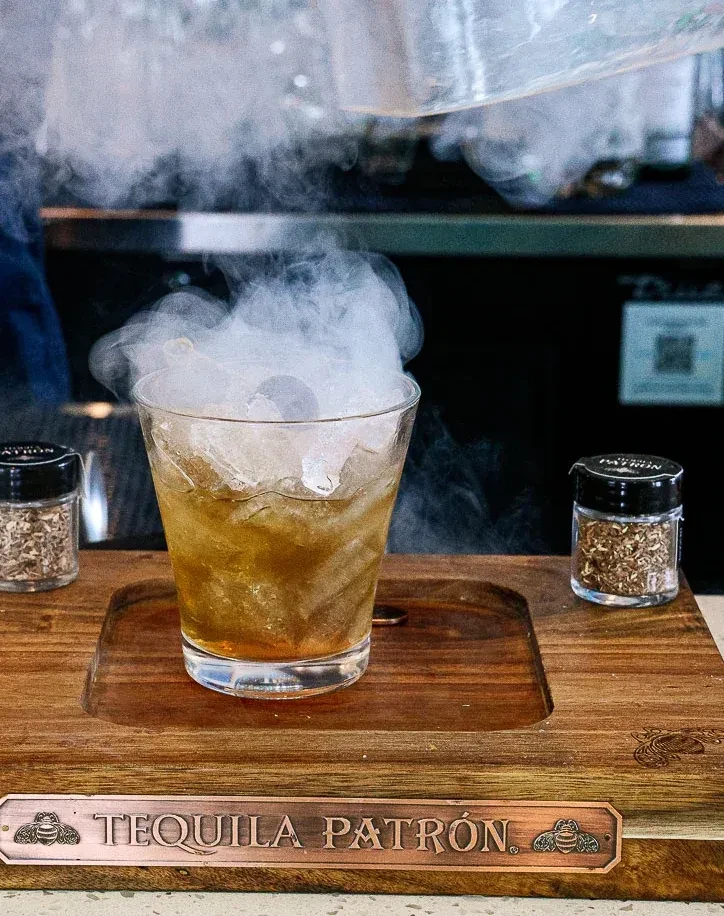 A smoky amber cocktail, Tequila Patrón plaque, and garnishes on tray.