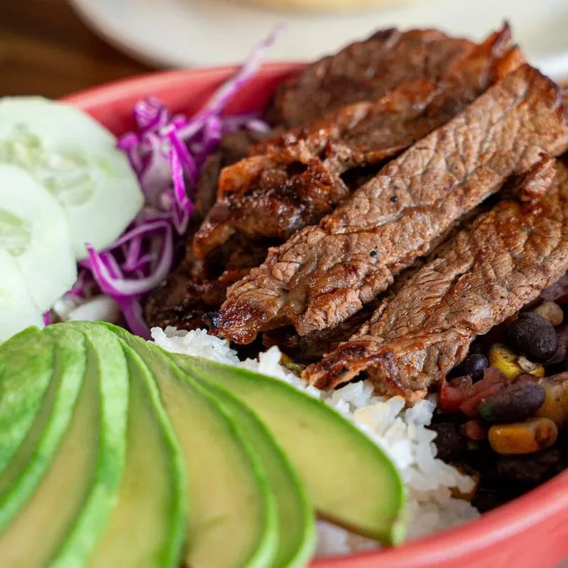 Red bowl with grilled steak, rice, black beans, corn, and vegetables.