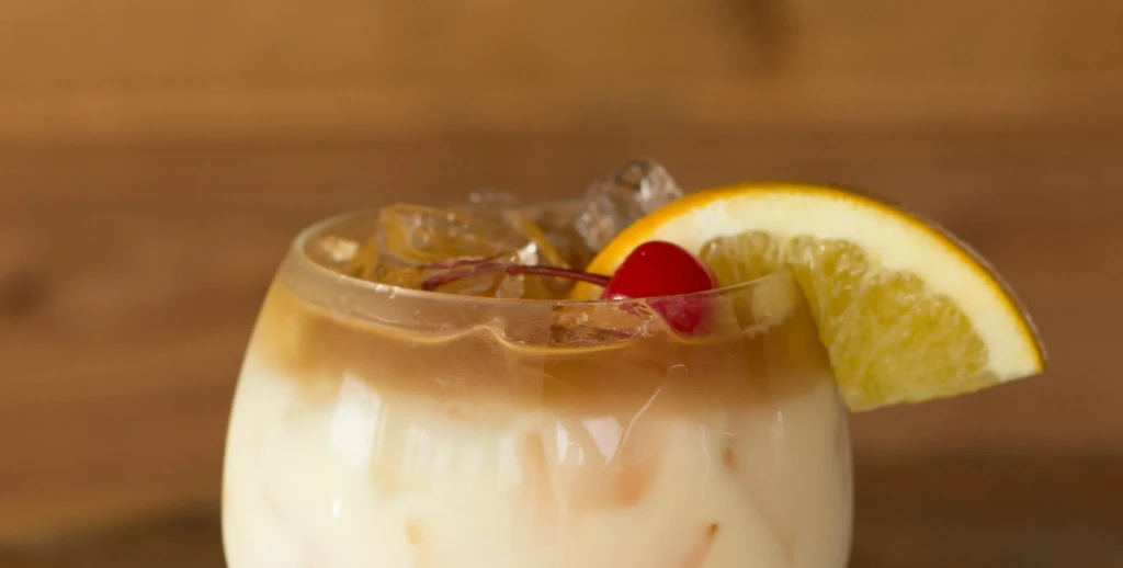 Layered creamy and amber drink with ice, cherry, and citrus garnish.