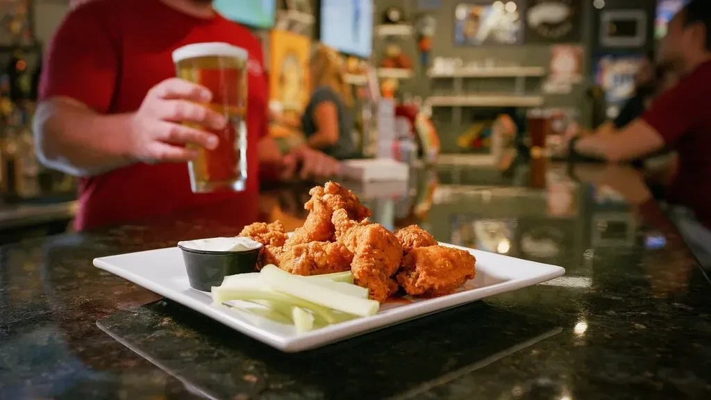 Fried chicken wings, celery, sauce, and beer on a bar counter.