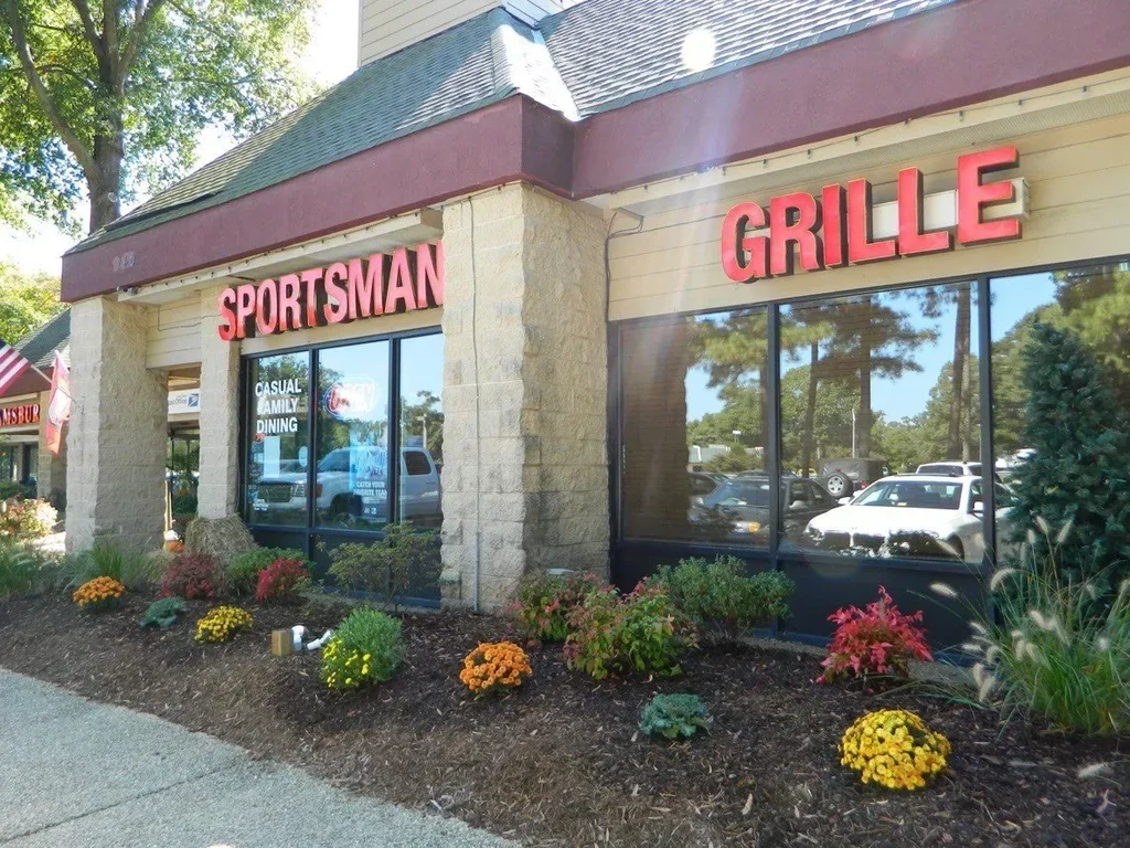 Sunny exterior of Sportsman Grille, stone pillars, red sign, colorful landscaping.