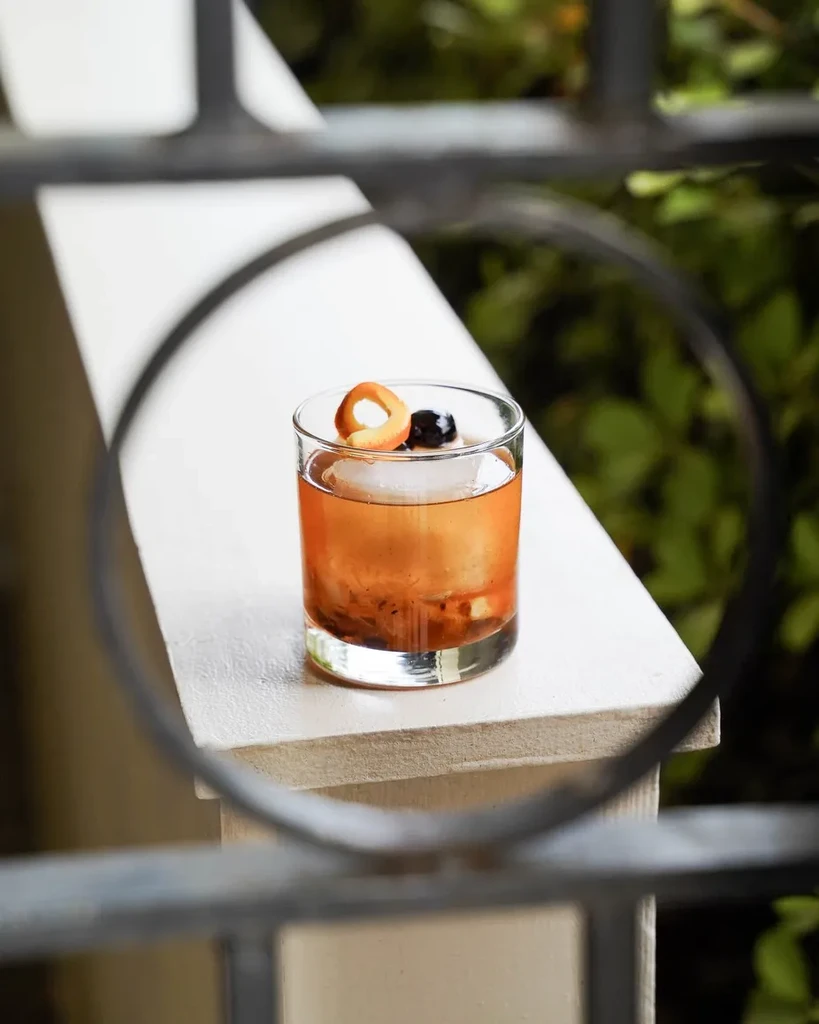 Reddish-brown cocktail with spherical ice and orange twist on outdoor ledge.