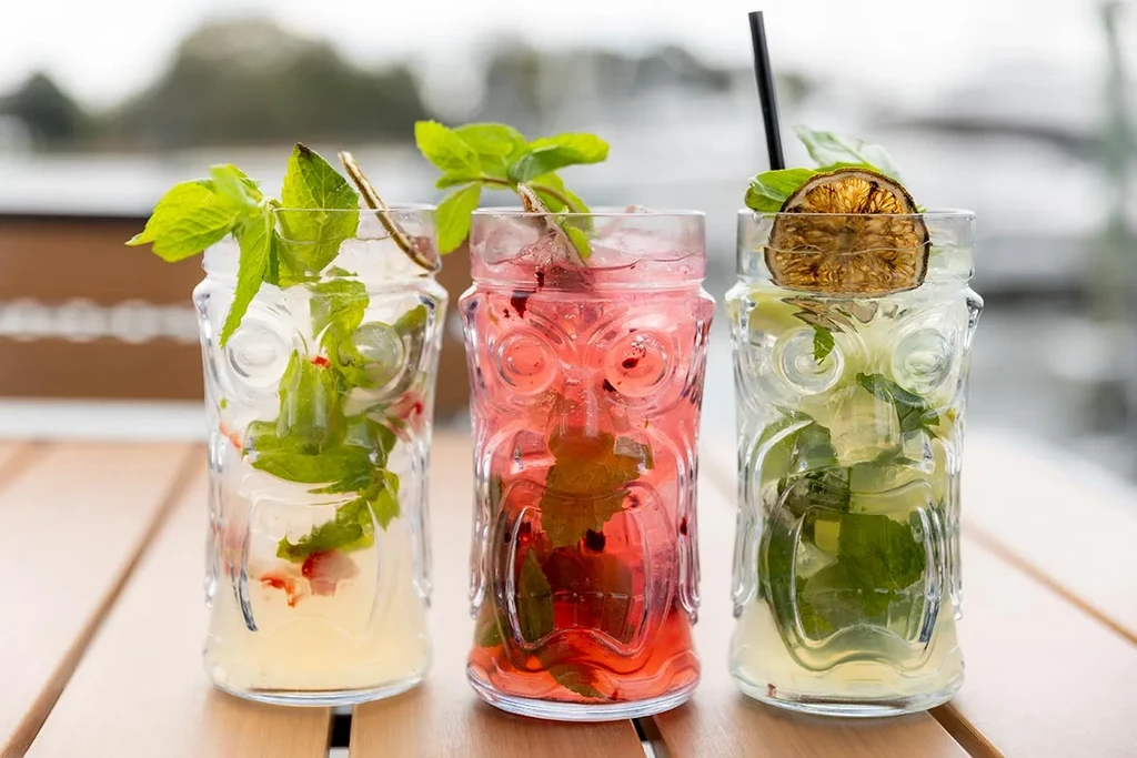 Three colorful cocktails in tiki head glasses, garnished with fruit and mint.