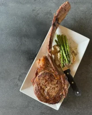 Tomahawk steak with asparagus and a creamy side on a white plate.