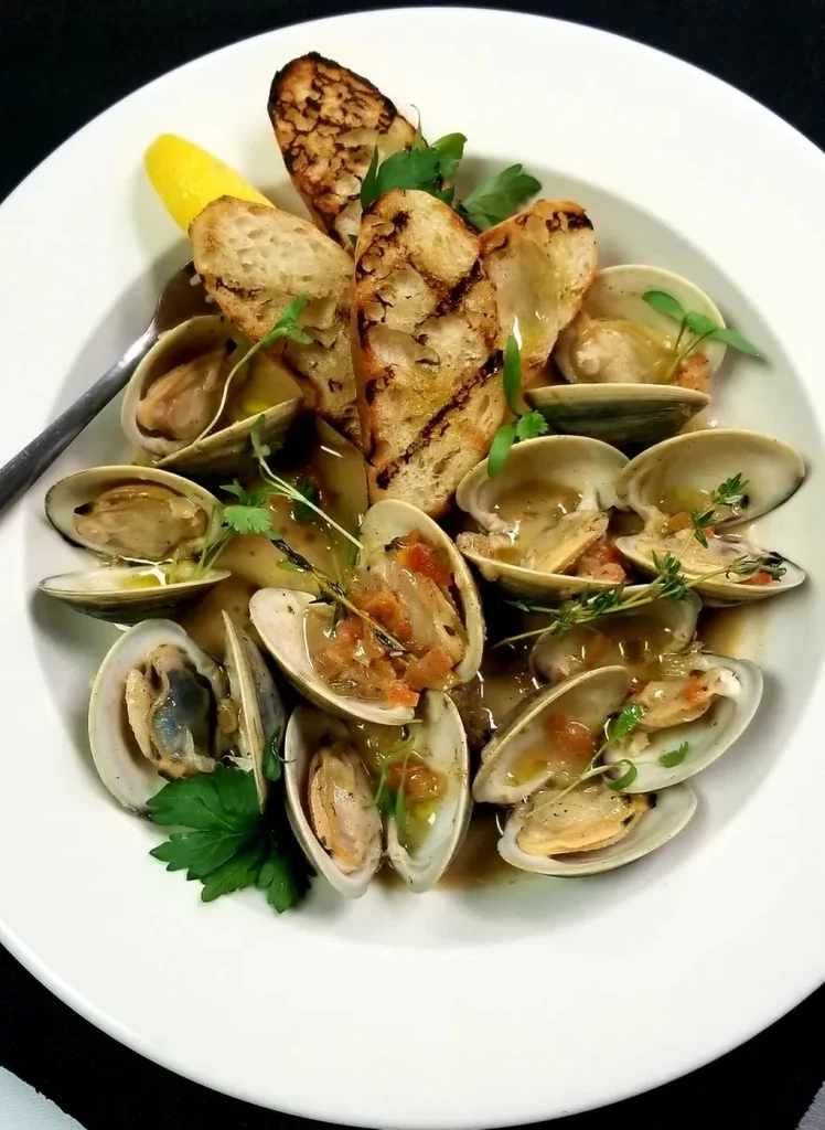 Steamed clams in a white bowl with baguette, lemon, and herbs.