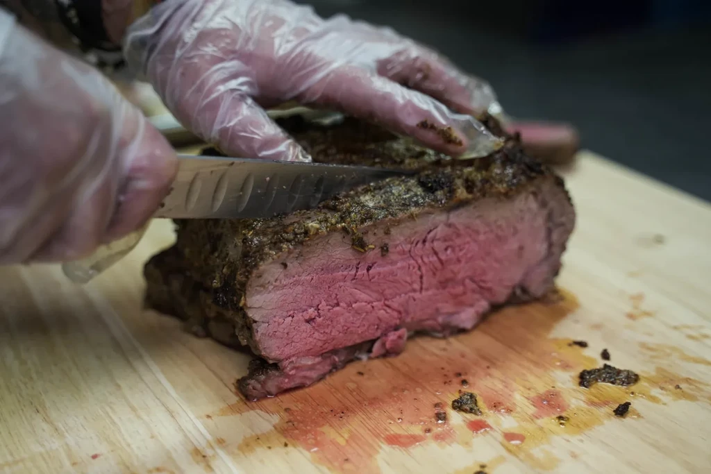 Hands in gloves slicing crispy, medium-rare, herb-crusted beef.