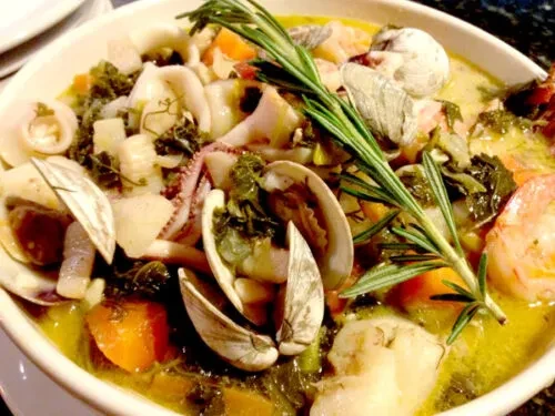 White bowl of seafood stew with shrimp, clams, vegetables, and rosemary.