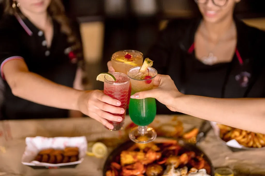 Three hands toast colorful cocktails with seafood and fries.