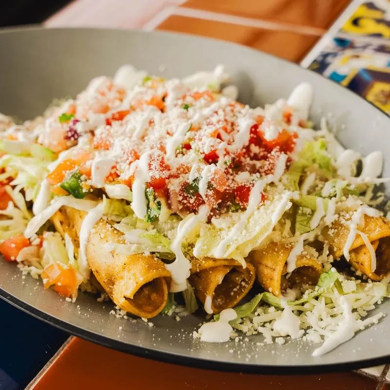 Close-up of golden taquitos topped with lettuce, tomatoes, cheese, and cream.