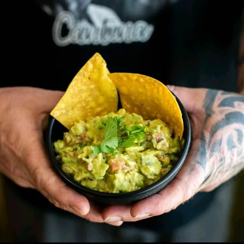 Tattooed hands hold chunky green guacamole with chips and cilantro.