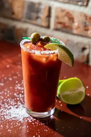 Red cocktail with salted rim, ice, green olives, and lime wedge.