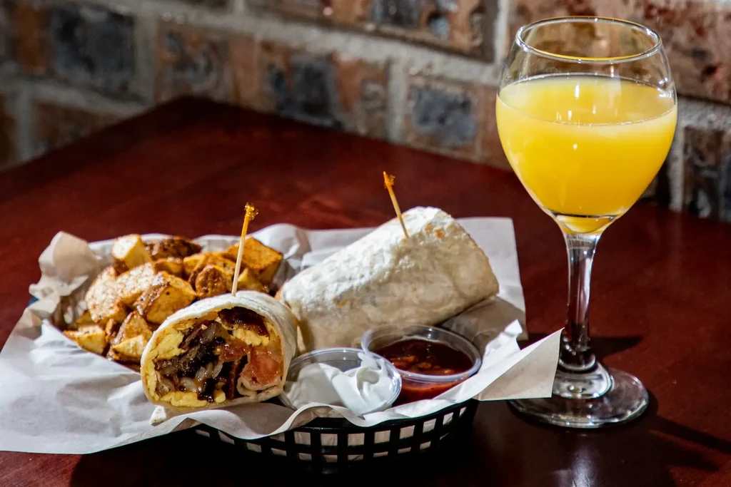 Two breakfast burritos, roasted potatoes, sauces, and orange juice on a table.