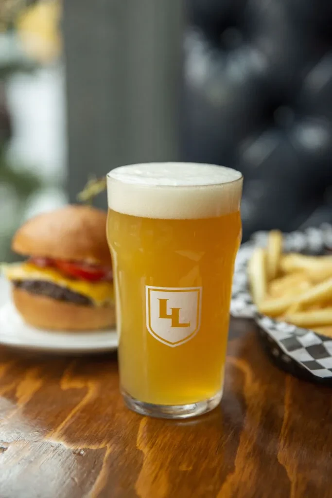 Frothy amber beer glass with LL logo, cheeseburger, and fries.