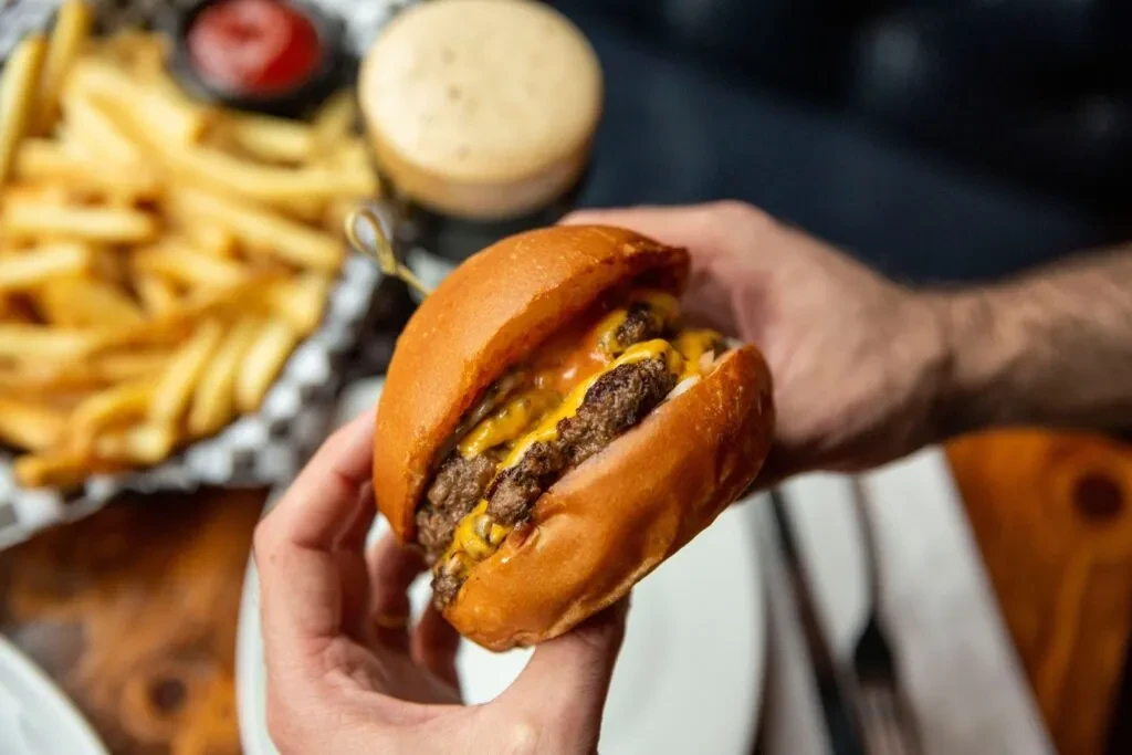 Two hands hold a double cheeseburger with melted cheese and a skewer.