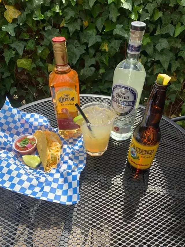 Two hard-shell tacos, salsa, margarita, beer, and tequila bottles on a patio.
