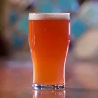 Pint glass of beer with frothy head on a reflective wooden surface.