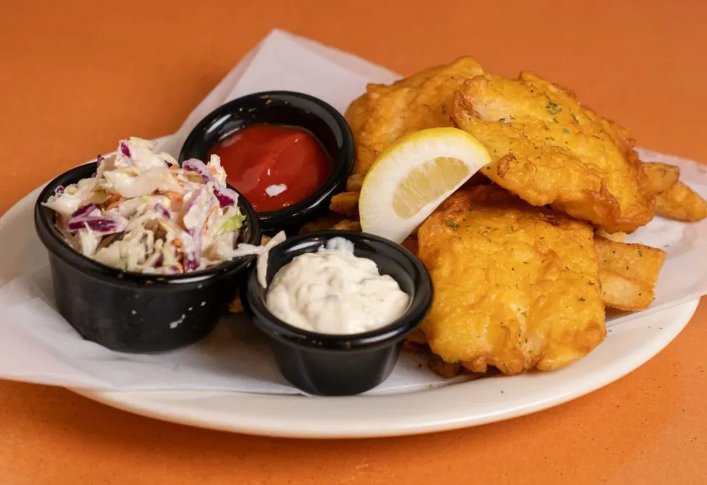 Golden-brown fish and chips with coleslaw, ketchup, tartar, and lemon wedge.
