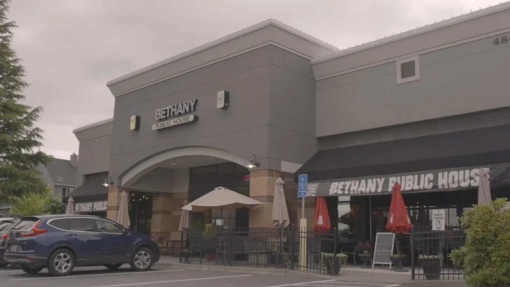 Gray Bethany Public House exterior with awning, outdoor seating, and parked vehicles.