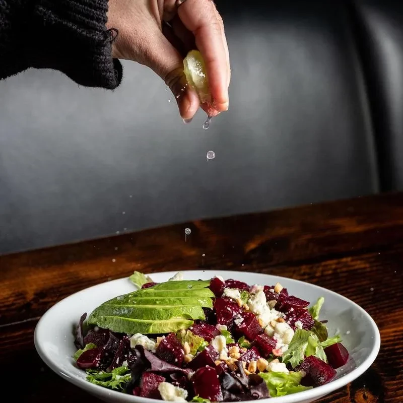 Hand squeezes citrus over a vibrant salad with avocado, beets, and cheese.