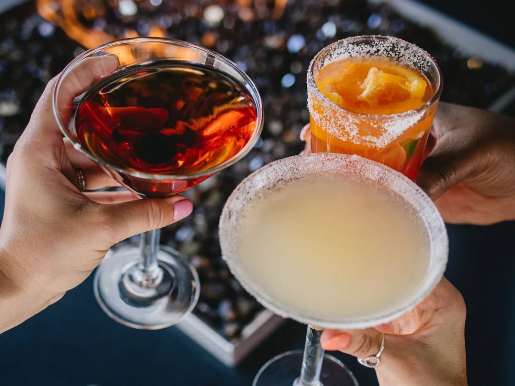 Three hands clink colorful cocktails against a glowing fire pit.