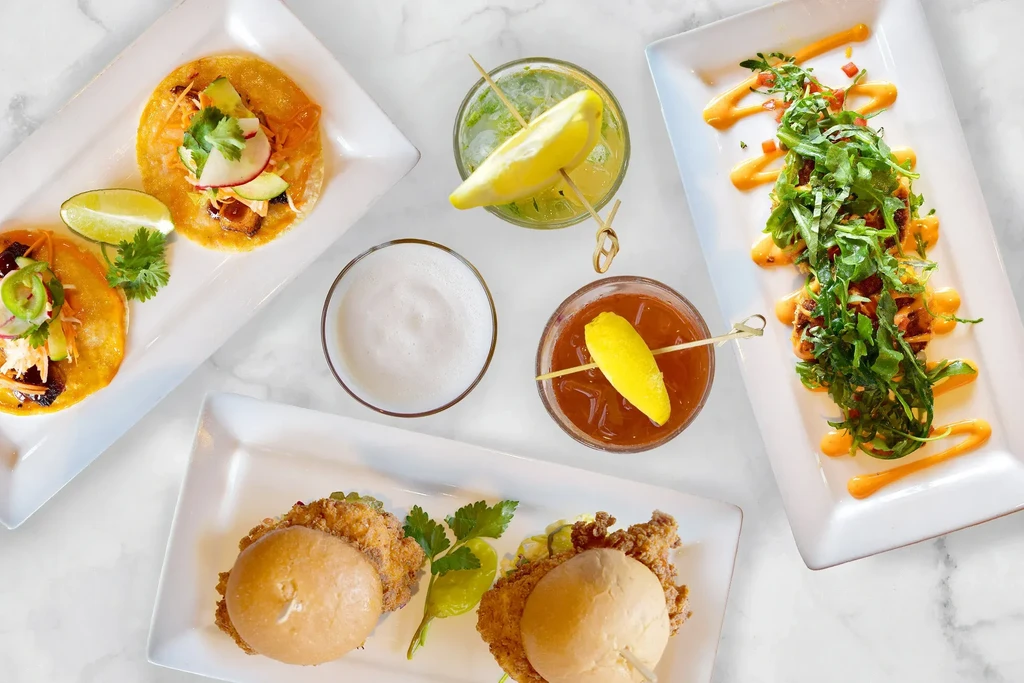 Overhead view of tacos, sliders, salad, and three beverages on marble.
