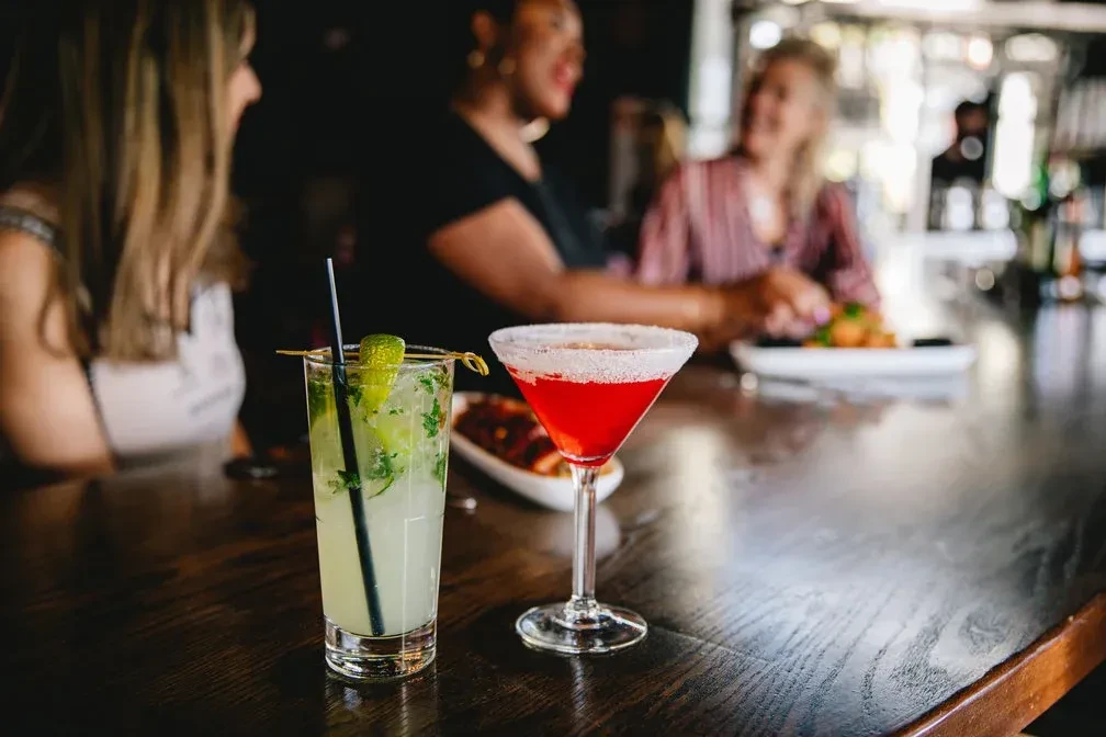 Green and red cocktails on a bar with blurred women talking behind.