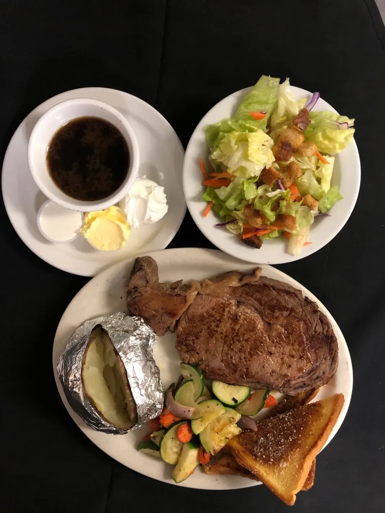 Multi-plate steak dinner with potato, vegetables, salad, and condiments.