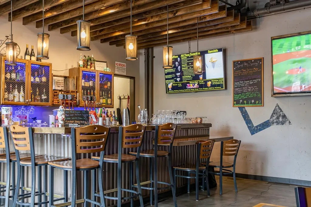Industrial bar with blue-lit bottles, digital menus, and baseball game.