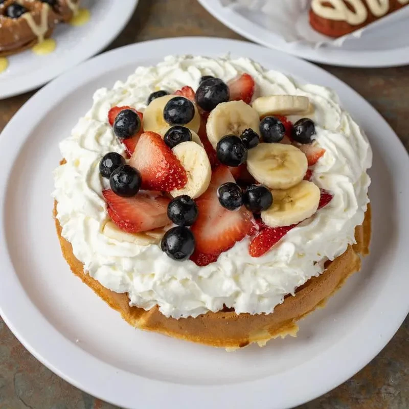 Waffle covered in whipped cream, strawberries, bananas, and blueberries on a plate.