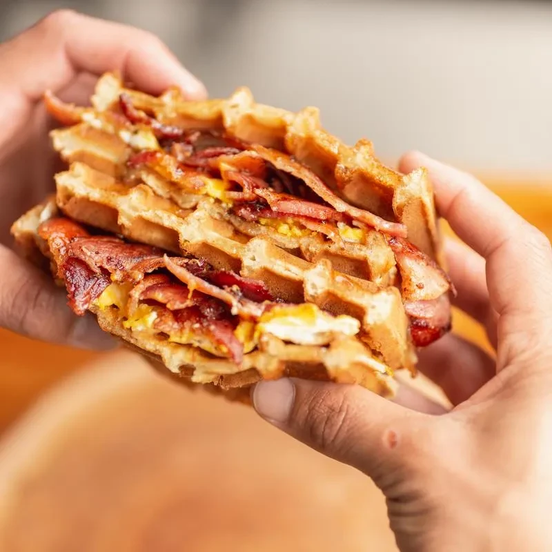 Close-up of hands holding a crispy waffle sandwich with bacon and eggs.