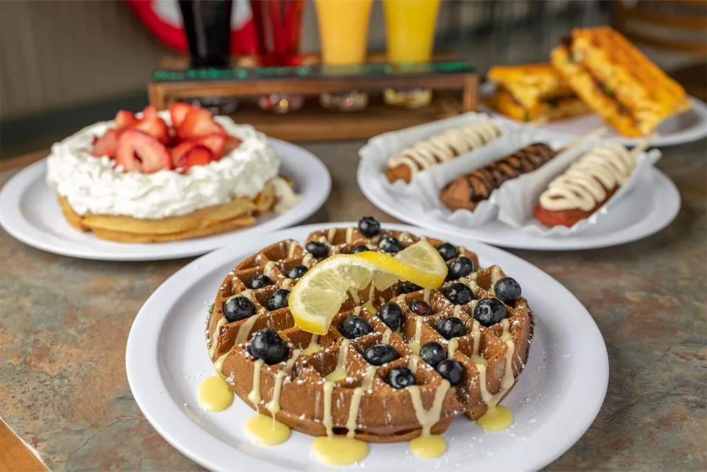 A breakfast and dessert spread of waffles, churros, sandwiches, and drinks.