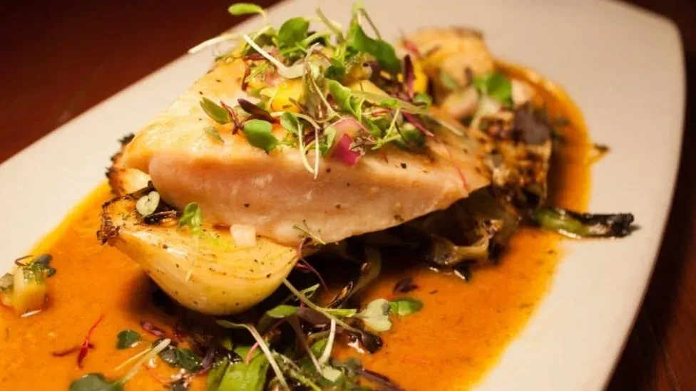 Plated fish fillet, microgreens, and orange-brown sauce on a modern white plate.