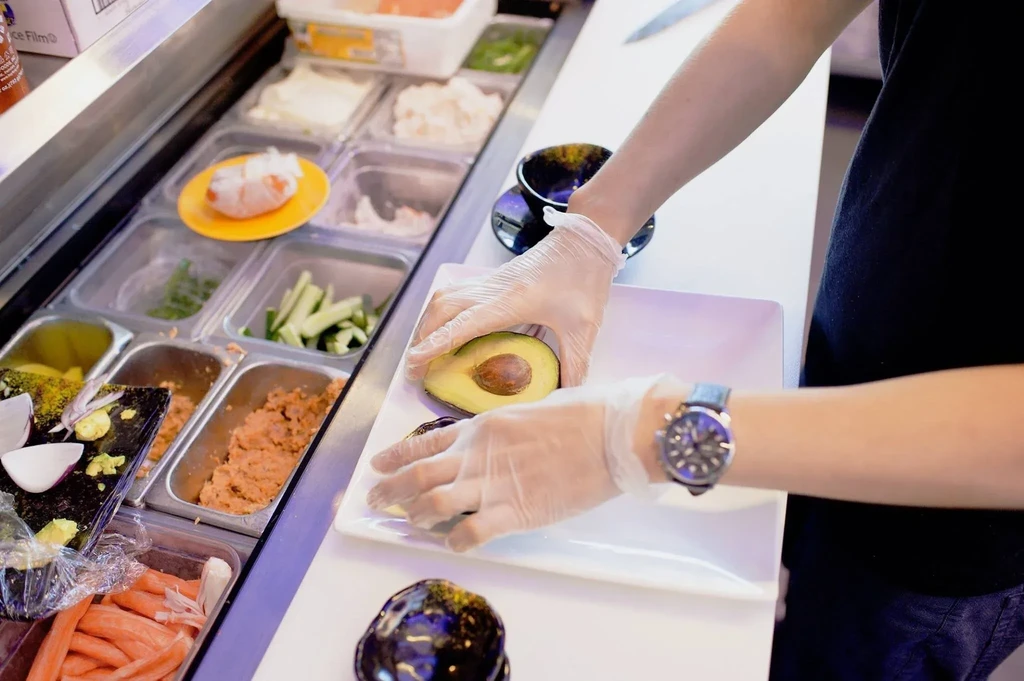 Gloved hands arranging a halved avocado on a plate at a food counter.