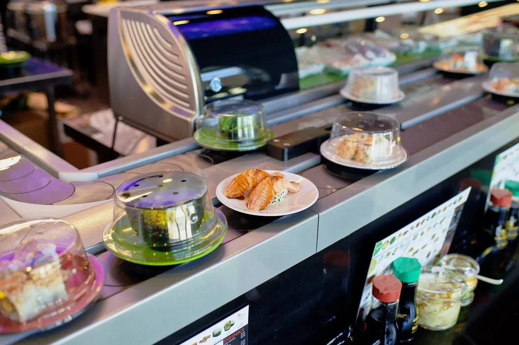 Seared salmon nigiri on a sushi conveyor belt with condiments nearby.