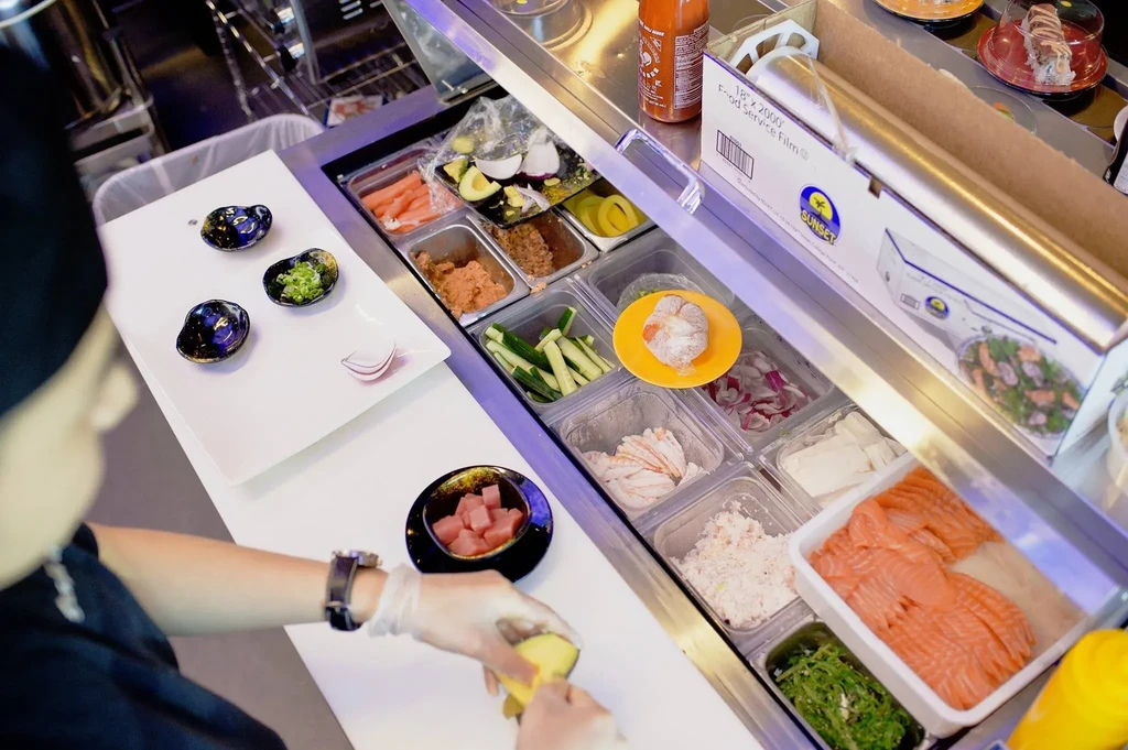 Chef's gloved hands peel avocado at a fresh sushi/poke ingredient display.