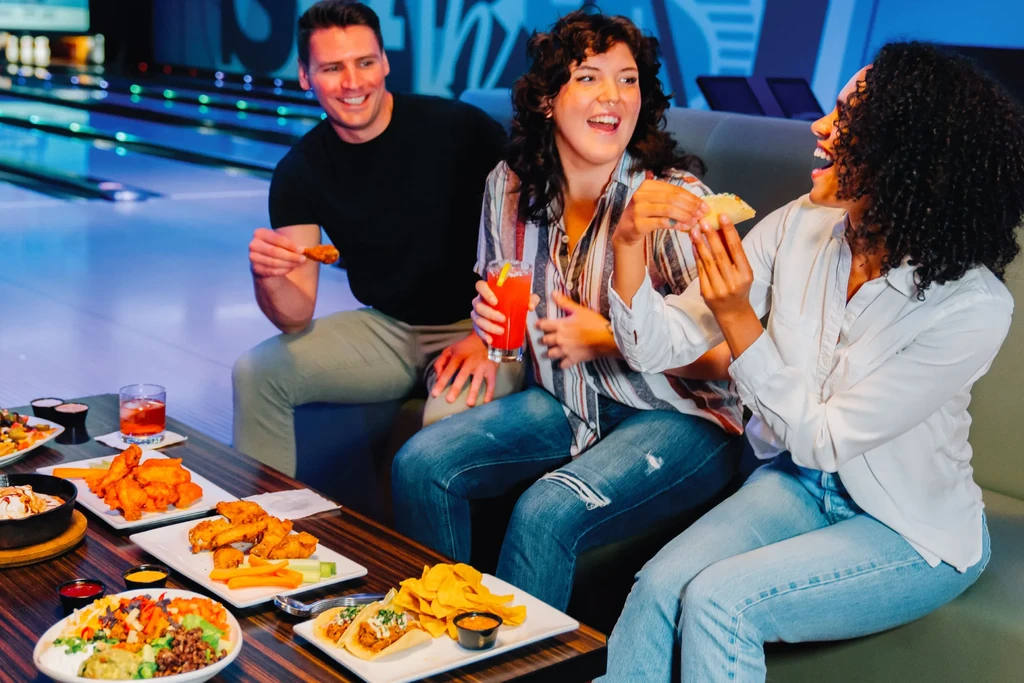 Smiling group eats wings and tacos in a neon-lit bowling alley.