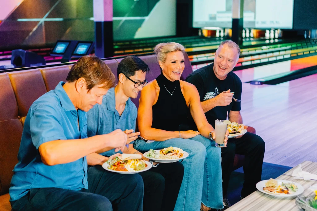 Four adults eating and laughing in a bowling alley booth.