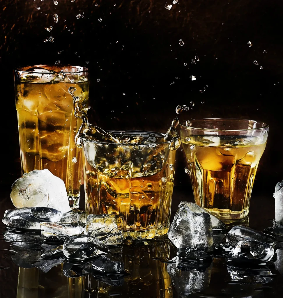 Glasses of amber liquid with ice, one splashing on dark background.