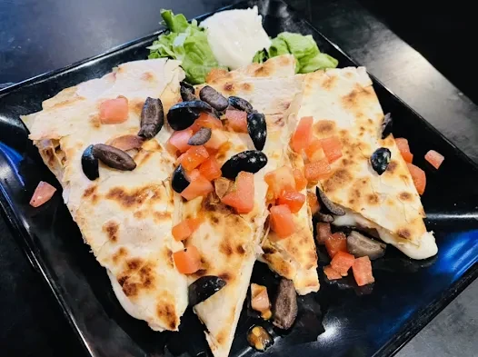 Golden-brown quesadilla wedges with tomatoes, olives, and cheese on black plate.